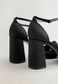 Sorte højhælede sandaler med mat finish, der har en bred blokhæle og en justerbar ankelrem. Ribbede detaljer på overdelen.