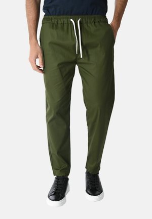 Uomo con pantaloni verde oliva con coulisse, scarpe da ginnastica nere con suola bianca e camicia nera con le mani nelle tasche.