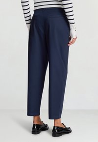 Pantaloni sartoriali blu navy con una vestibilità rilassata, caratterizzati da due tasche posteriori e una texture liscia. Indossati con mocassini neri.