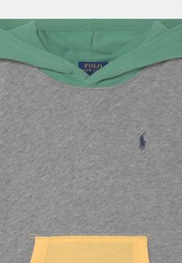 Harmaa collegepaita vihreällä hupulla ja keltaisella taskulla. Rinnan kohdalla pieni laivastonsininen logo ja pehmeä kangaspinta.