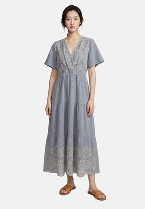 Femme portant une robe bleu-gris clair à encolure en V avec broderie florale et manches courtes, associée à des sandales plates couleur tabac, debout sur un fond clair.