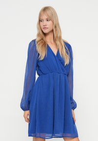 Tânără cu părul lung blond, îmbrăcată cu o rochie albastră regală până la genunchi, cu mâneci lungi transparente și decolteu în V, stând într-un fundal simplu.