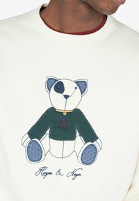 Bluza ma kremowe tło z kolorowym, haftowanym wzorem misia. Miś ma niebieskie uszy, zielony sweter i akcenty w postaci guziczków.