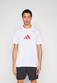 Valge puuvillane T-särk, mille ees on suur punane kolmnurkne graafiline logo ja tekst. Lühikesed varrukad ja ümmarguse kaelusega disain.