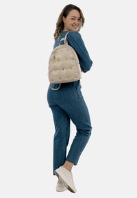 Bees padded seljakott kuldsete detailidega ja nittakestega, millel on monogrammikas. Kandnud mudel, kes on riietatud teksadega.