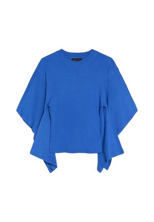Femme en haut bleu et pantalon foncé ajustant de grandes lunettes de soleil noires, debout devant de hauts bâtiments en verre réfléchissant.
