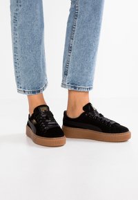 Svarta sammets sneakers med en tjock brun gummisul, med platta svarta snören och en subtil guld logotyp på tungan.