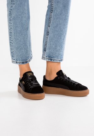 Zapatillas de terciopelo negro con una suela gruesa de goma marrón, con cordones planos negros y un sutil logo dorado en la lengüeta.