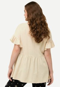 Blusa beige chiaro con maniche corte a volant, vita leggermente peplum e una texture liscia. Indossata con pantaloni neri a fantasia con forme geometriche.