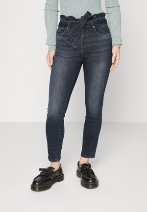 Femme portant un jean en denim bleu foncé taille haute avec une taille de type paperbag et une ceinture nouée, associé à des mocassins à franges noirs, se tenant sur un sol clair.