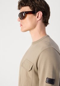 Sweatshirt bege com um bolso no peito e gola canelada; possui um logo na manga e uma textura de tecido suave. Óculos de sol escuros incluídos.