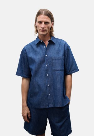 Homme aux cheveux mi-longs portant une chemise bleue à manches courtes boutonnée et un short bleu assorti sur un fond uni.