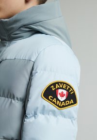 Doudoune bleu clair avec capuche, ornée d'une bande en tissu sur la manche gauche affichant "ZAVETTI CANADA" et une feuille d'érable rouge.