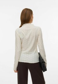 Top met lange mouwen in crème, met een delicaat patroon met kleine donkere accenten. Getailleerd model met een opstaande kraag, gedragen met donkere broek.