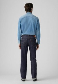 Camisa de mezclilla azul claro de manga larga, combinada con jeans azul oscuro que cuentan con un parche de cuero y detalles de costura visibles.