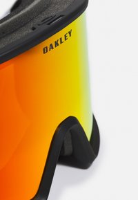 Lunettes Oakley avec une monture noire et des verres dégradés allant du orange au jaune. Les verres présentent un revêtement réfléchissant et des bords texturés pour une meilleure prise.