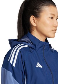 Marineblauem, wasserdichtem Jacke mit Kapuze, featuring weißen Streifen an den Schultern und einem Reißverschluss. Glatte Textur und sportliches Design.