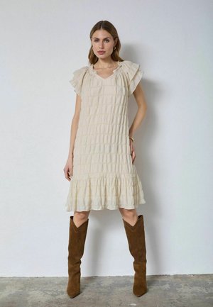 Žena oblečená v krémovom textúrovanom šatách do kolien s volánmi na rukávoch a lemoch, v kombinácii s vysokými hnedými semišovými topánkami, stojaca pri bielej stene.