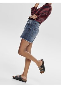 Jeans-Minirock mit fransigem Saum, leicht bis mittelblau gewaschen, und zwei hinteren Taschen. Getragen mit einem rot gestreiften Langarmoberteil und schwarzen Sandalen.