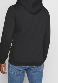 Sudadera con capucha negra hecha de una tela suave, con un bolsillo tipo canguro y puños acanalados, combinada con jeans de un azul claro.