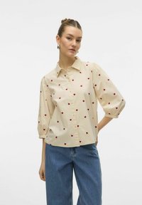 Beige knäppt skjorta med korta puffärmar, prydd med små röda hjärtmönster. Tillverkad av ett lätt tyg. Bärs med blå jeans.