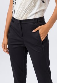 Pantalons extensibles noirs avec poches zippées sur les côtés, texture lisse et coupe ajustée. La ceinture a des passants pour une ceinture.
