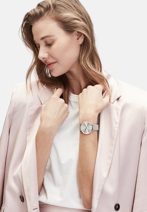 Silberne Uhr mit rundem Zifferblatt, grauem Armband und minimalistischem Design. Am Handgelenk getragen, ergänzt durch einen hellrosa Blazer und ein weißes Hemd.