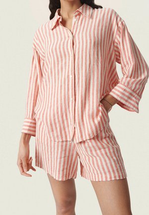 Mujer con camisa de manga larga de rayas verticales en rosa y blanco holgada y shorts a juego, con una mano en el bolsillo.
