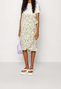 Wit T-shirt gecombineerd met een gele wikkelrok met bloemenprint en ruches, aangevuld met een lavendel tas en witte sandalen met plateauzool.