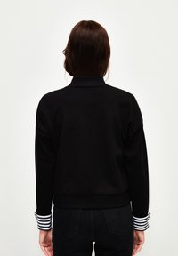 Schwarzer Sweatshirt mit hohem Kragen und gestreiften Bündchen. Hat eine lockere Passform und einen gerippten Saum. Glatte Textur, minimale Details.