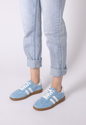 Hellblaue Wildledersneaker mit weißen Streifen und Schnürsenkeln, ausgestattet mit einer braunen Gummisohle und einem glatten weißen Kragen. Die Jeans sind an den Knöcheln hochgekrempelt.