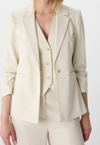 Blazer beige clair avec revers crantés, fermeture à deux boutons et manches longues, superposé sur un gilet sans manches assorti. Tissu texturé.