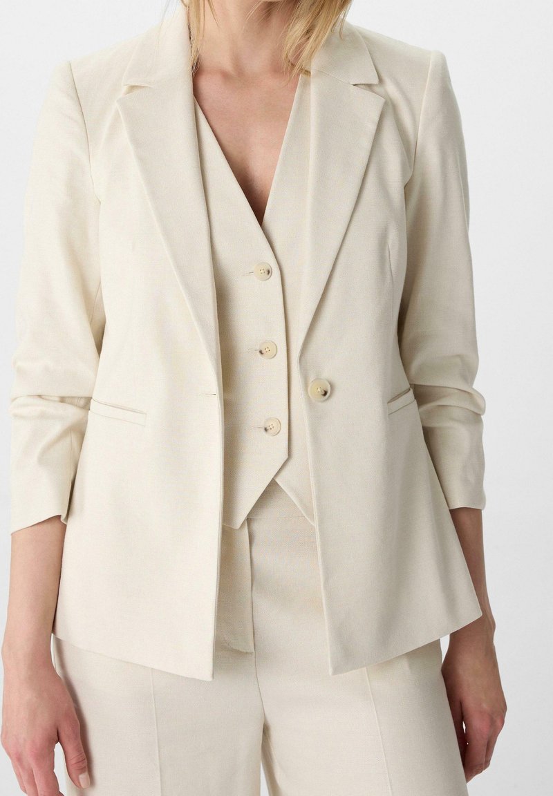 Blazer beige clair avec revers crantés, fermeture à deux boutons et manches longues, superposé sur un gilet sans manches assorti. Tissu texturé.