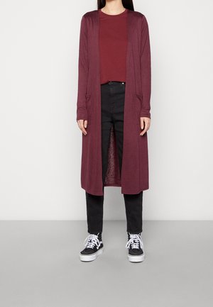 Personne portant un long cardigan bordeaux sur une chemise bordeaux assortie, un pantalon noir et des baskets noires et blanches sur un fond uni.