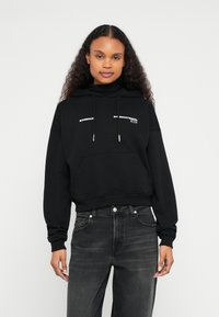 Camisola de capuz preta cropped de algodão com bolso frontal e cordões. Apresenta logótipo em texto branco no peito. Combinada com jeans escuros.