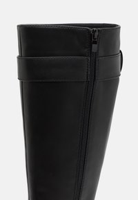 Bota de cuero negro con un diseño elegante, que cuenta con una cremallera lateral, costuras verticales y un empeine texturizado con un borde superior redondeado.