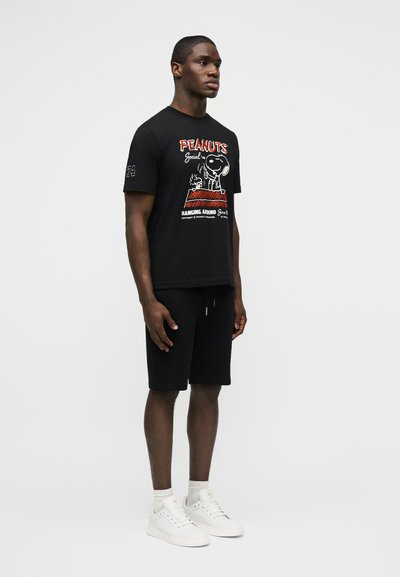 Άνδρας που στέκεται φορώντας μαύρο t-shirt με το λογότυπο Peanuts, μαύρες βερμούδες, λευκές κάλτσες και λευκά αθλητικά παπούτσια, σε απλό φωτεινό φόντο.