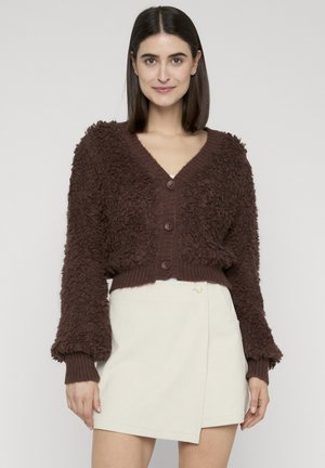 Kvinna som bär en texturerad brun kofta med knappar och en vit asymmetrisk kjol, stående mot en enfärgad ljus bakgrund.