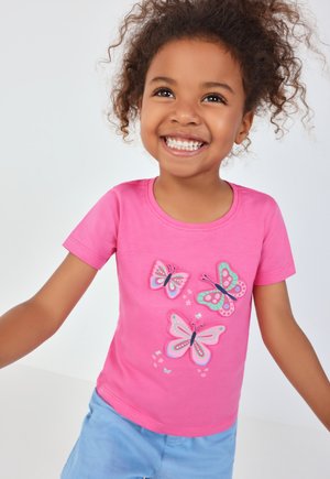 Glimlachend jong meisje in roze t-shirt met kleurrijke vlinderprints en lichtblauwe shorts, poserend met uitgestrekte armen tegen een effen achtergrond.