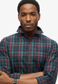 Geruit overhemd in donker groen, blauw en bordeauxrood met een knoopsluiting. Heeft een klassieke kraag en een stoftextuur met een klein logodetail.