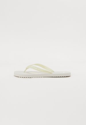 Chanclas blancas con tiras amarillas claras, que cuentan con una suela texturizada y el nombre de la marca impreso en la tira. Diseño simple y perfil plano.