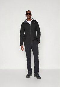 Black lightweight jacket with a zip front, gray accents, paired with dark pants and sturdy black hiking shoes. Model wearing sunglasses.