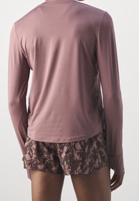 Langärmliges, mauvefarbenes Sportoberteil mit glatter Textur, kombiniert mit gemusterten braunen Shorts mit floralem Design und gerafftem Saum.