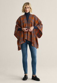 Orangeternet plaid poncho med brede ærmer og åben front, båret over en navy turtleneck og jeans, med et blødt, tekstureret stof.