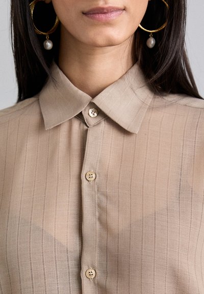 Camicia button-up beige a righe con colletto, realizzata in tessuto traslucido e con bottoni beige. Il modello indossa orecchini a cerchio con un accento di perla.