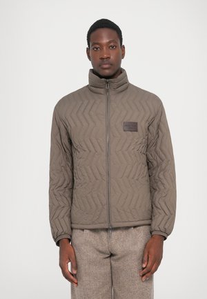 Olive green quilted jacket with high collar, front zipper, and textured chevron pattern. Features a small branded patch on left chest.