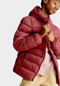 Ung person iført maroon pufferjakke, som de zipper op, med beige bukser og en lys pink skjorte synlig nedenunder.