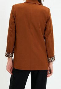 Blazer marrón con solapa de muesca, mangas largas y puños de estampado de leopardo. Tejido suave con una forma estructurada y ajuste entallado.