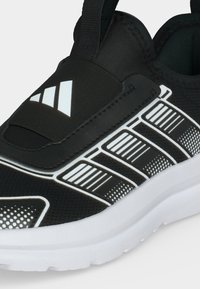 Μαύρο αθλητικό παπούτσι τύπου slip-on με άσπρη σόλα, με άσπρα ριγέ και πουά σχέδια και άσπλο τριγωνικό λογότυπο στο ελαστικό επάνω μέρος.