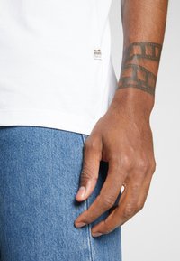 Weißes Baumwoll-T-Shirt mit kleinem G-Star Raw Logo am Saum, kombiniert mit blauen Jeans. Die Hand ruht lässig auf der Jeans.